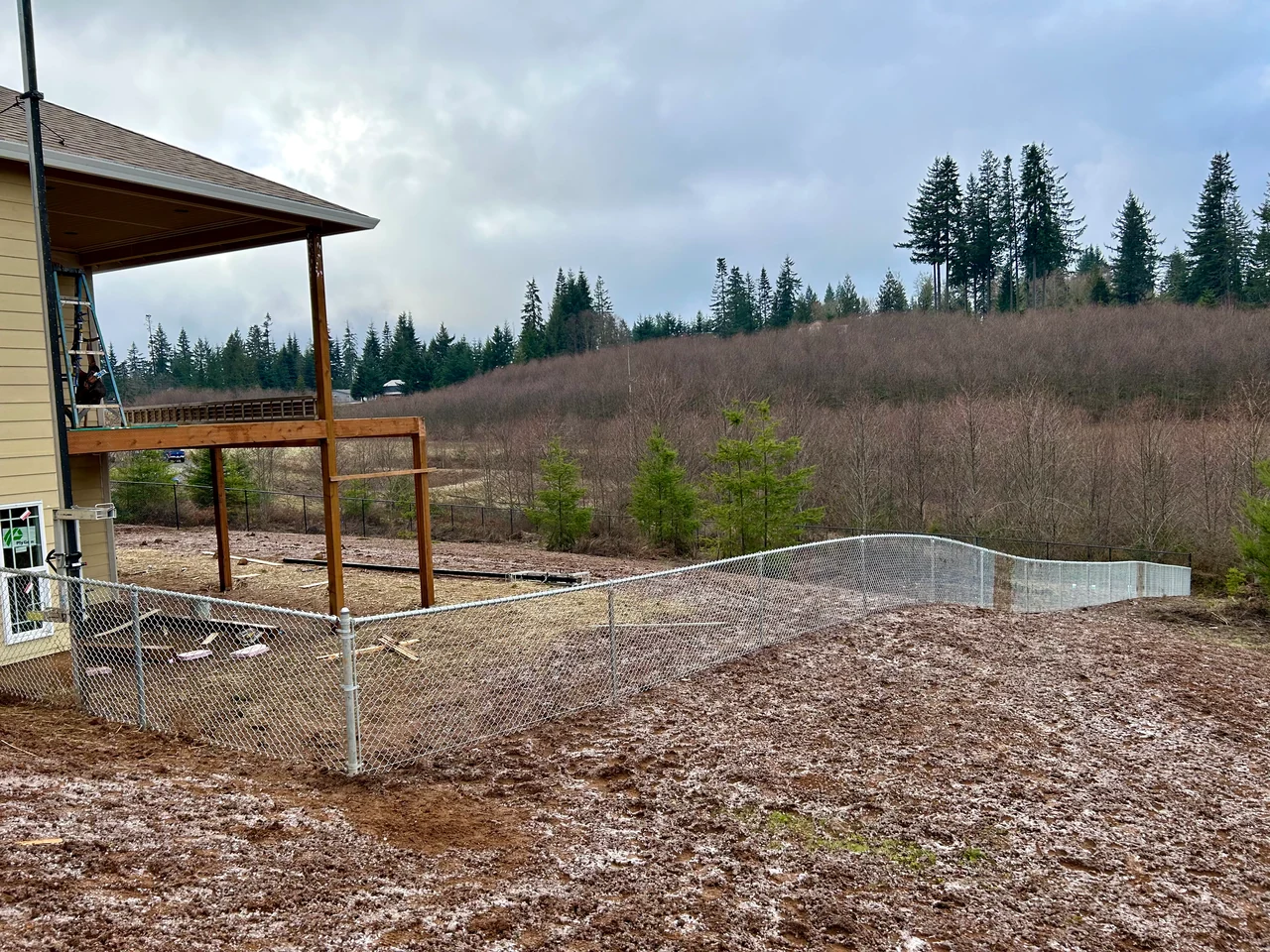 Silver chain link fencing follows a sloped yard beside a house with a raised covered deck and a wooded hillside beyond.