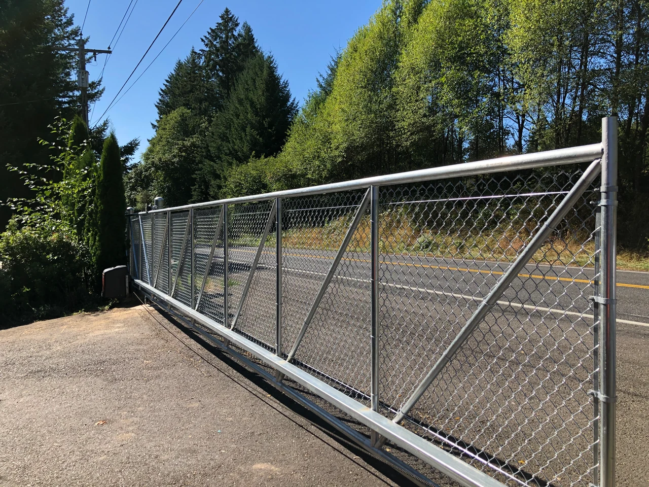 A wide chain link sliding gate with diagonal bracing spans a paved entrance beside trees and a street.