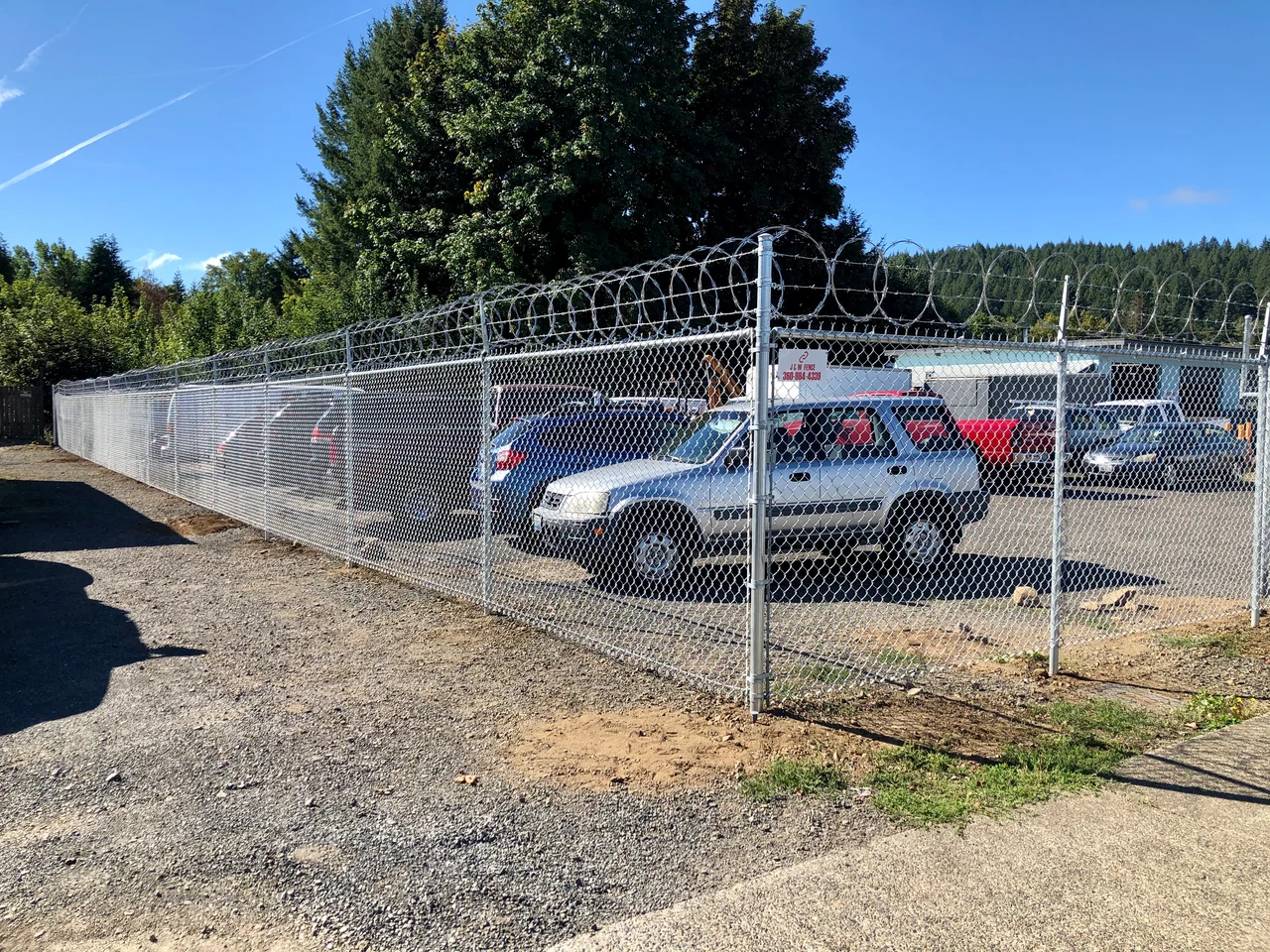 Silver chain link fencing topped with razor ribbon surrounds a vehicle lot, with a J & W Fence sign on the corner post.