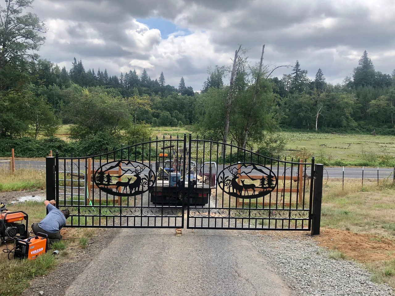 A worker fits a black double driveway gate with decorative wildlife panels at the entrance to a rural property.