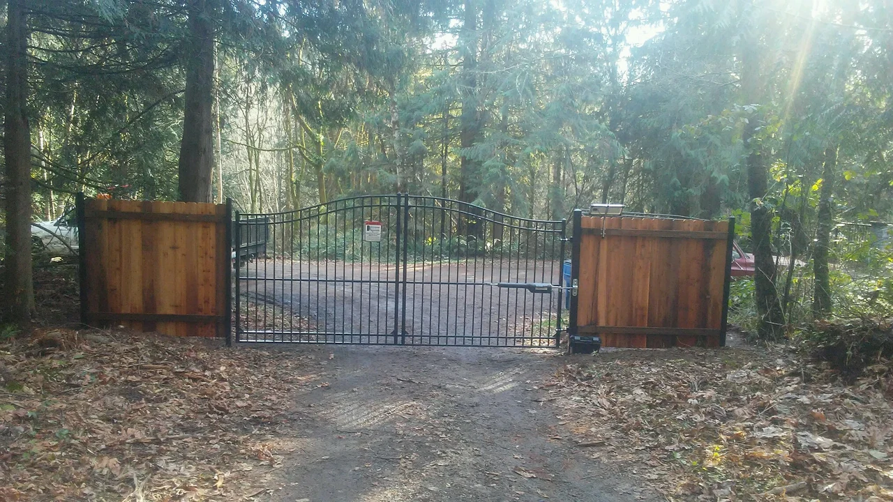 A black arched driveway gate stands between wood privacy panels at a shaded, tree-lined property entrance.