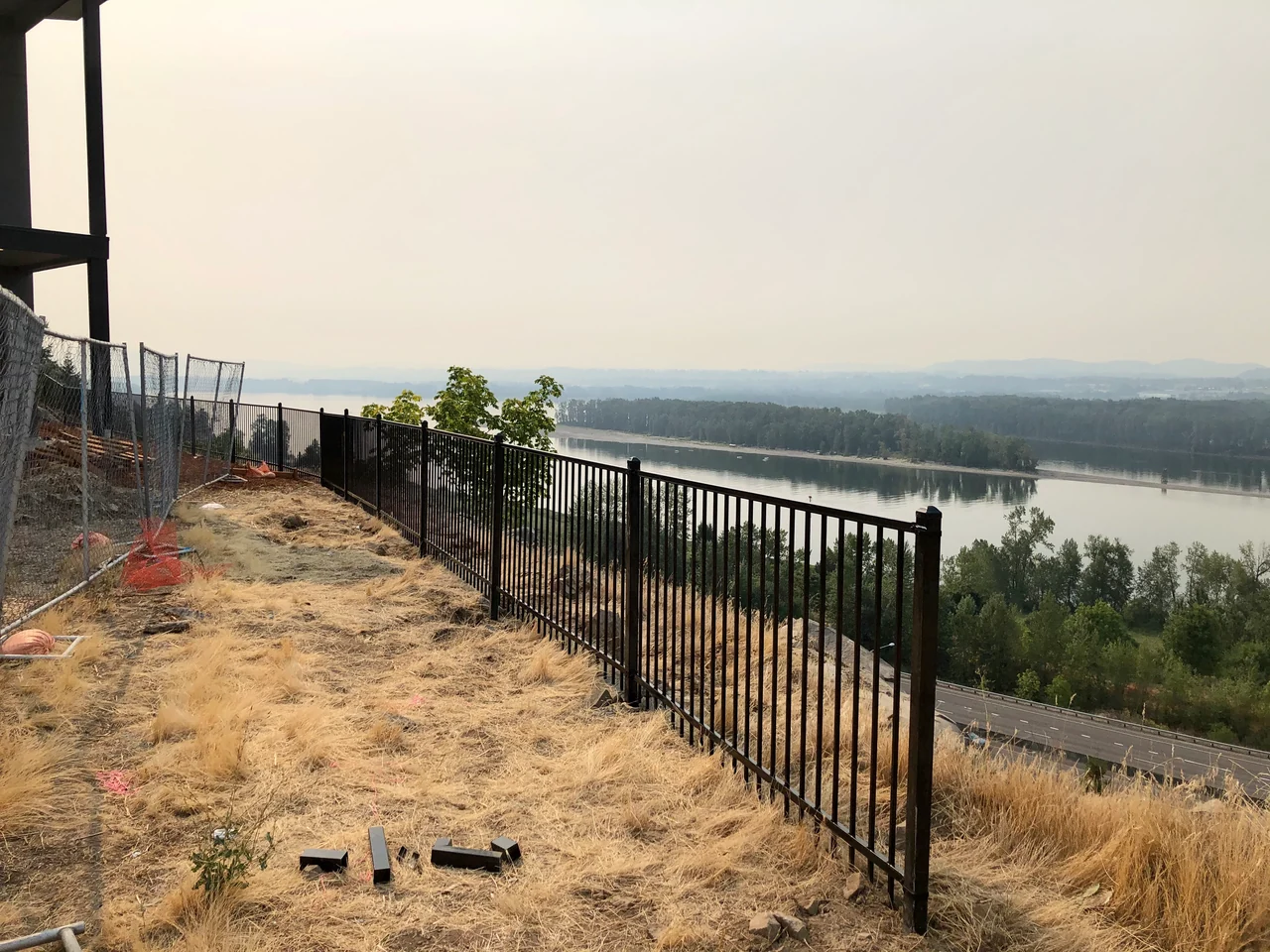 Black ornamental metal fencing follows a dry hillside overlooking a river, beside temporary construction fencing.