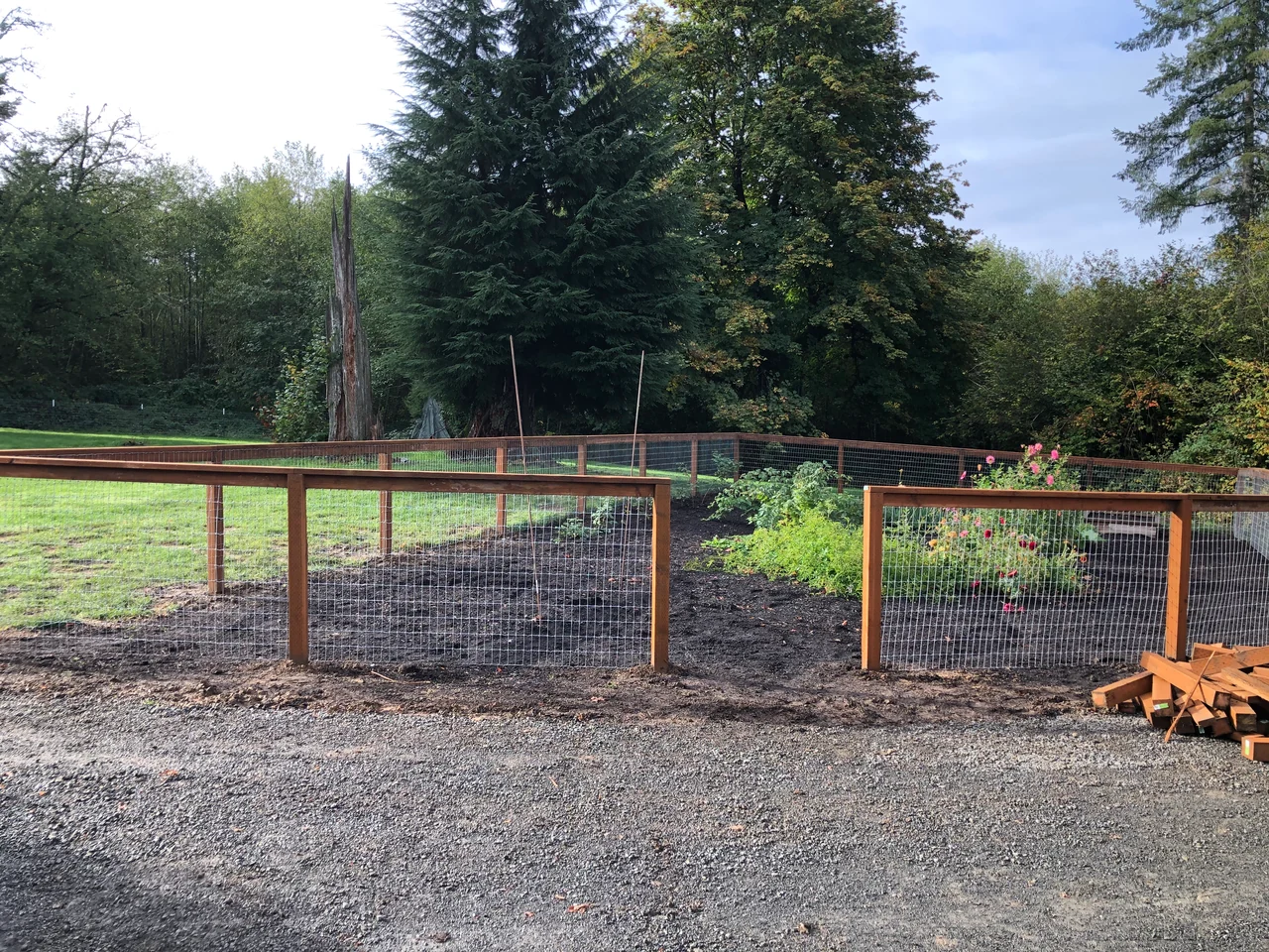 Wood posts and rails frame wire mesh around a garden and lawn, with an open entry and tall trees behind the enclosure.