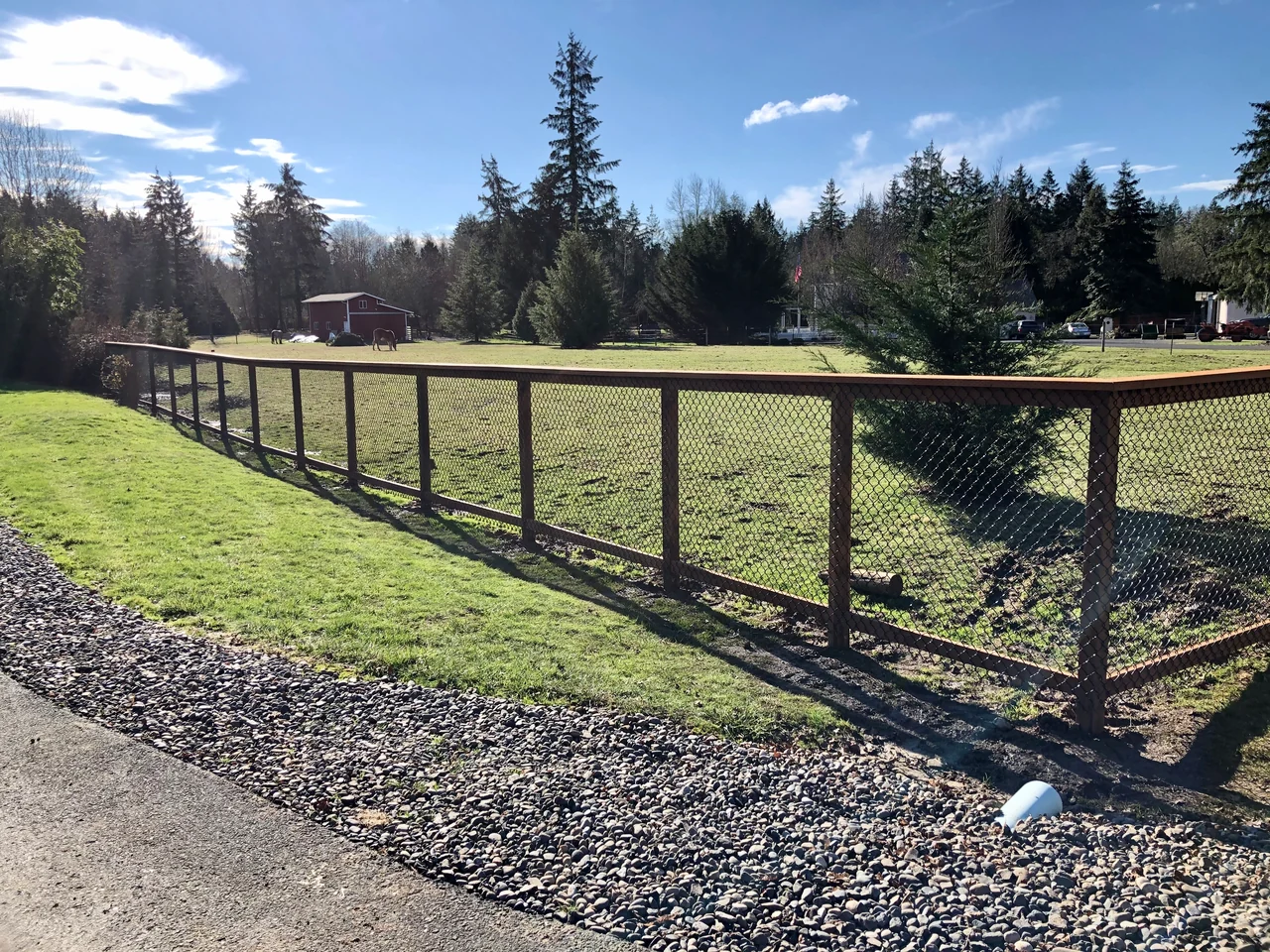 Wood posts and a top rail frame wire mesh fencing along a grassy field beside a gravel driveway.