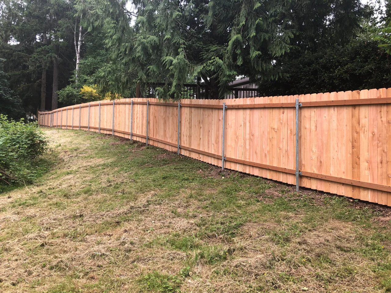 A long wood privacy fence with metal posts and horizontal support rails runs along a tree-lined lawn.