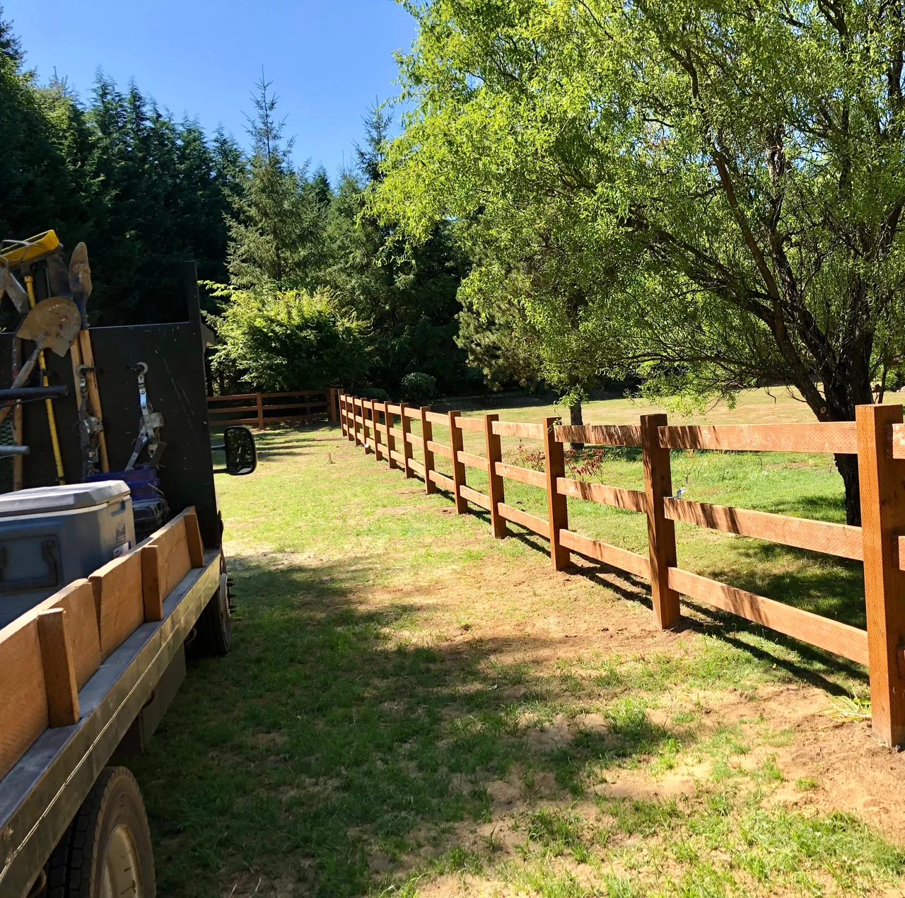 A wood three-rail fence runs beneath leafy trees alongside a grassy property, with a work truck at the edge of the view.